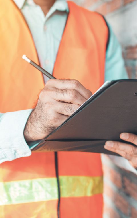 homen usando um colete laranja de trabalho segurando uma prancheta e escrevendo nela com lapis